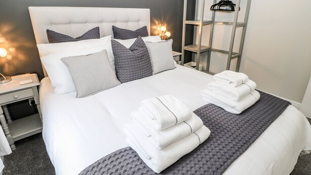 A neatly made bed with white linens and grey pillows, a grey textured rug, and a stack of white towels. - Victoria House (Holmfirth)