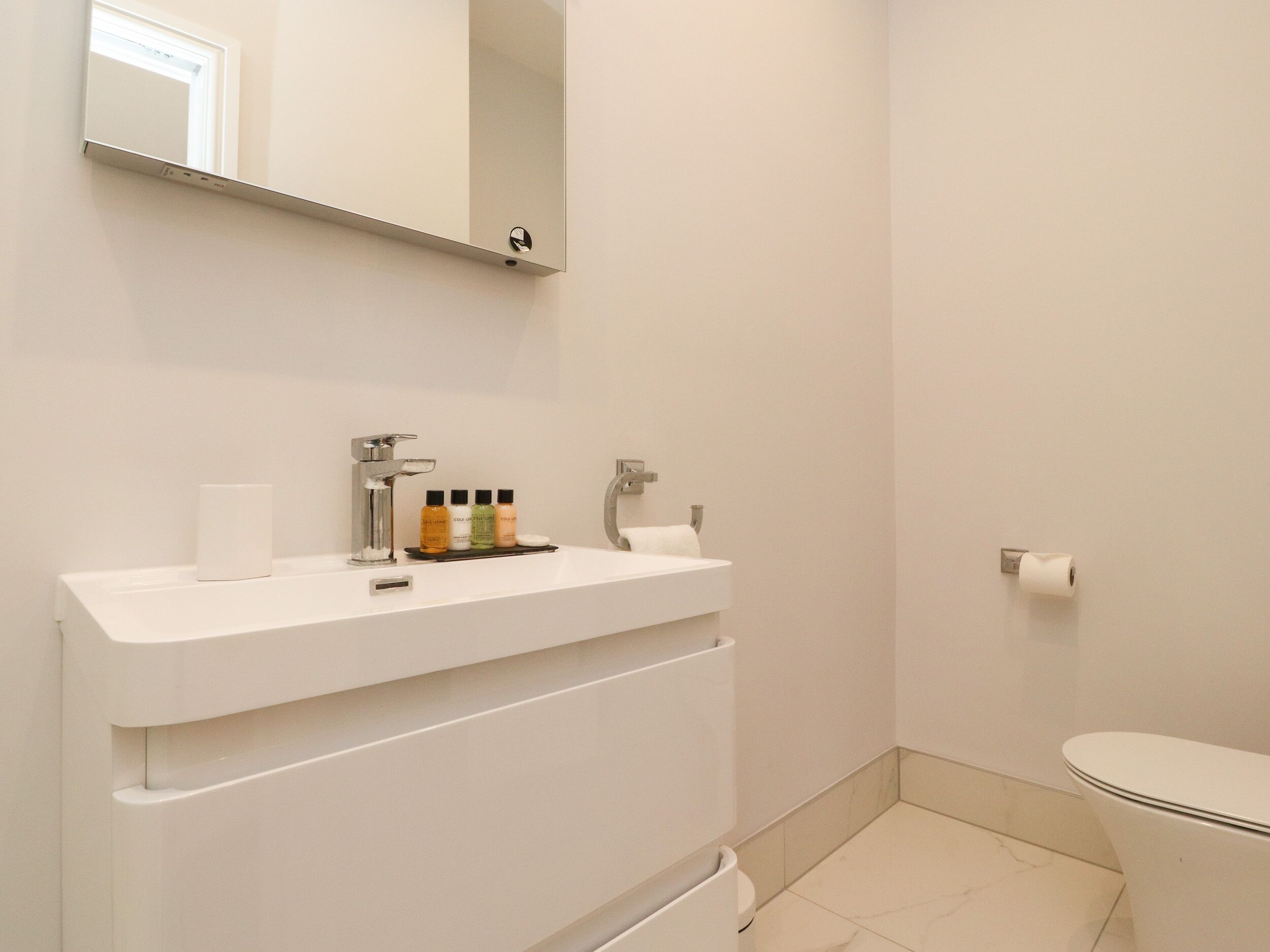 A bathroom with a white sink, mirror, and toilet. There are toiletries on the sink and a towel hanging on the wall.