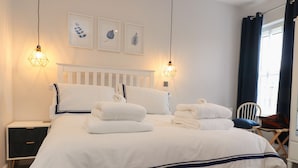 Una cama bien hecha con sábanas blancas y detalles en azul marino, una mesita de noche blanca con cajón, una obra de arte con hojas enmarcada en la pared y una ventana con cortinas oscuras.