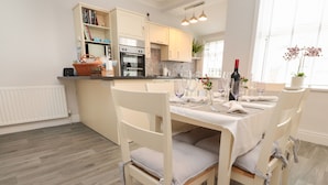 Una cocina moderna con armarios blancos, un comedor con mesa preparada para cuatro personas y una ventana con una orquídea en maceta.