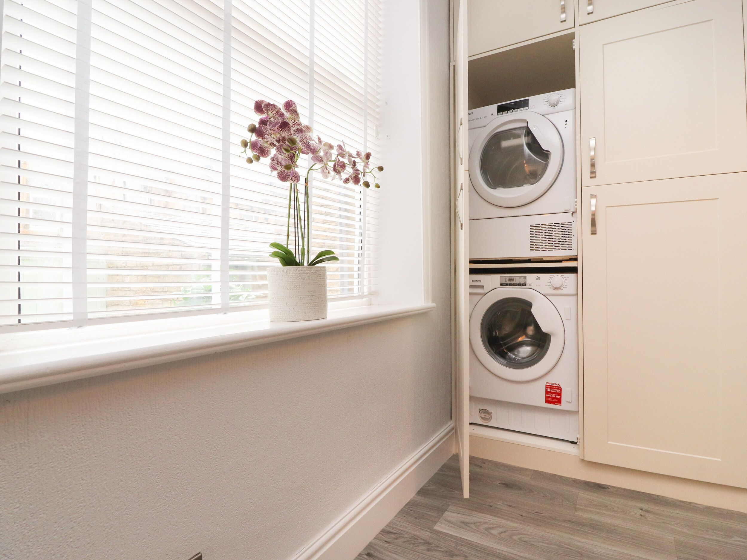 A laundry room with two stacked washing machines, a potted orchid on the windowsill, and white cabinetry.