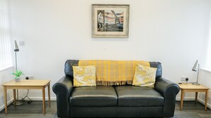 A living room with a black leather sofa, yellow throw, and floral cushions. There is a framed picture on the wall, a small table with a plant, and a patterned rug on the floor. - 2 Mountain View (Llangefni)
