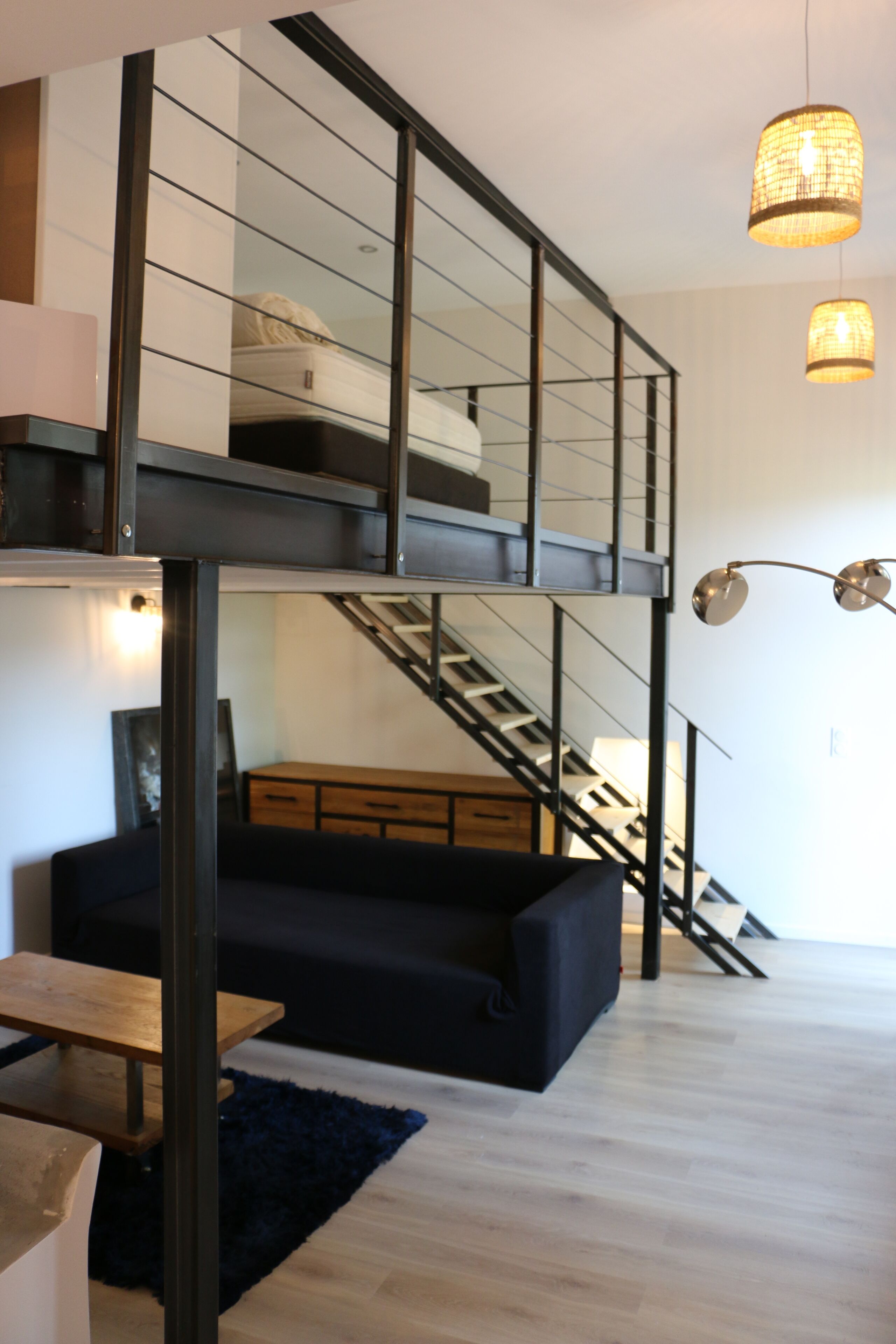 A modern loft-style living space with a high ceiling, a staircase, a sofa, a wooden table, and pendant lighting.