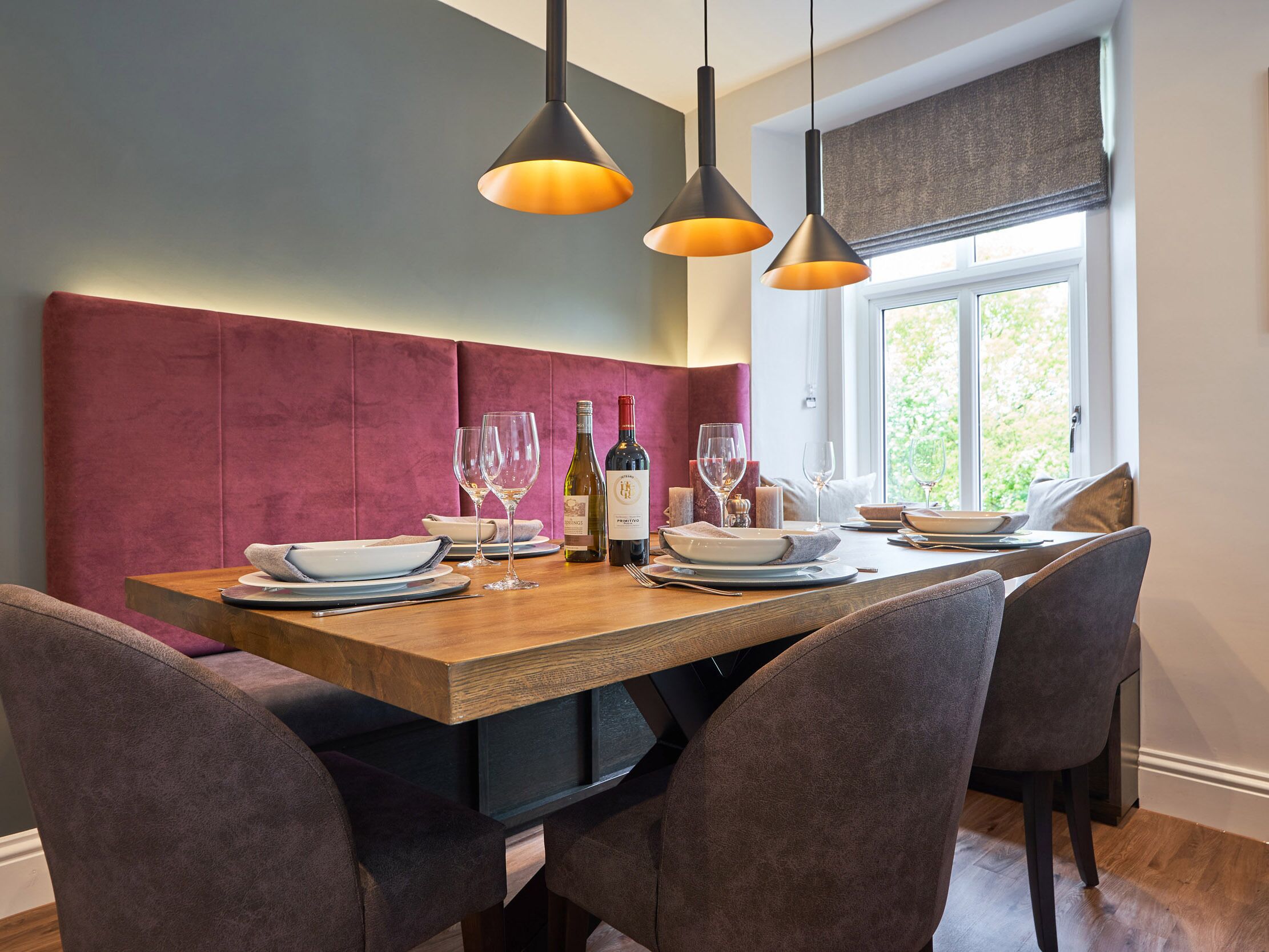 Un comedor con una mesa de madera dispuesta para cuatro comensales, dos lámparas colgantes y un ventanal con vista a zonas verdes.