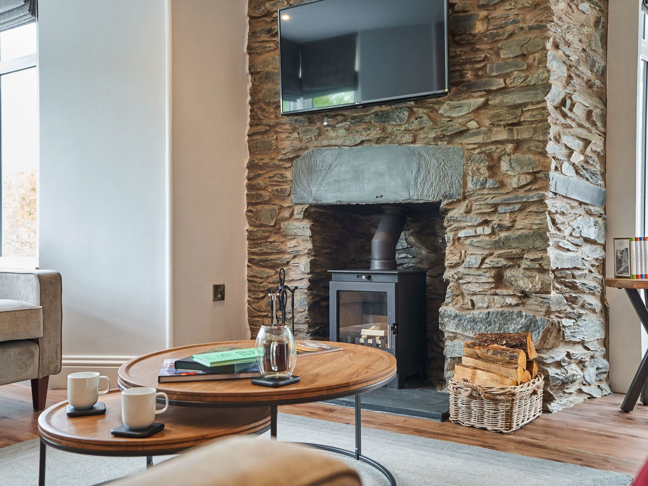 A cozy living room with a stone fireplace, a flat-screen TV mounted above it, and a round wooden table with two cups and a book.