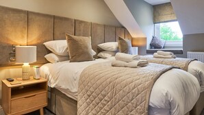A modern bedroom with two beds, a nightstand with a lamp, and a view of the outdoors through a window. - Upper Sheriff's Place (Windermere)