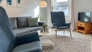 A modern living room with a grey sofa, a blue armchair, a wooden TV stand, and a large window with curtains. - Sünnslag Wohnung 064 (Ostseebad Boltenhagen)
