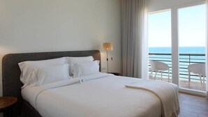 A hotel room with a large bed, a nightstand, a lamp, and a view of the sea through a balcony.