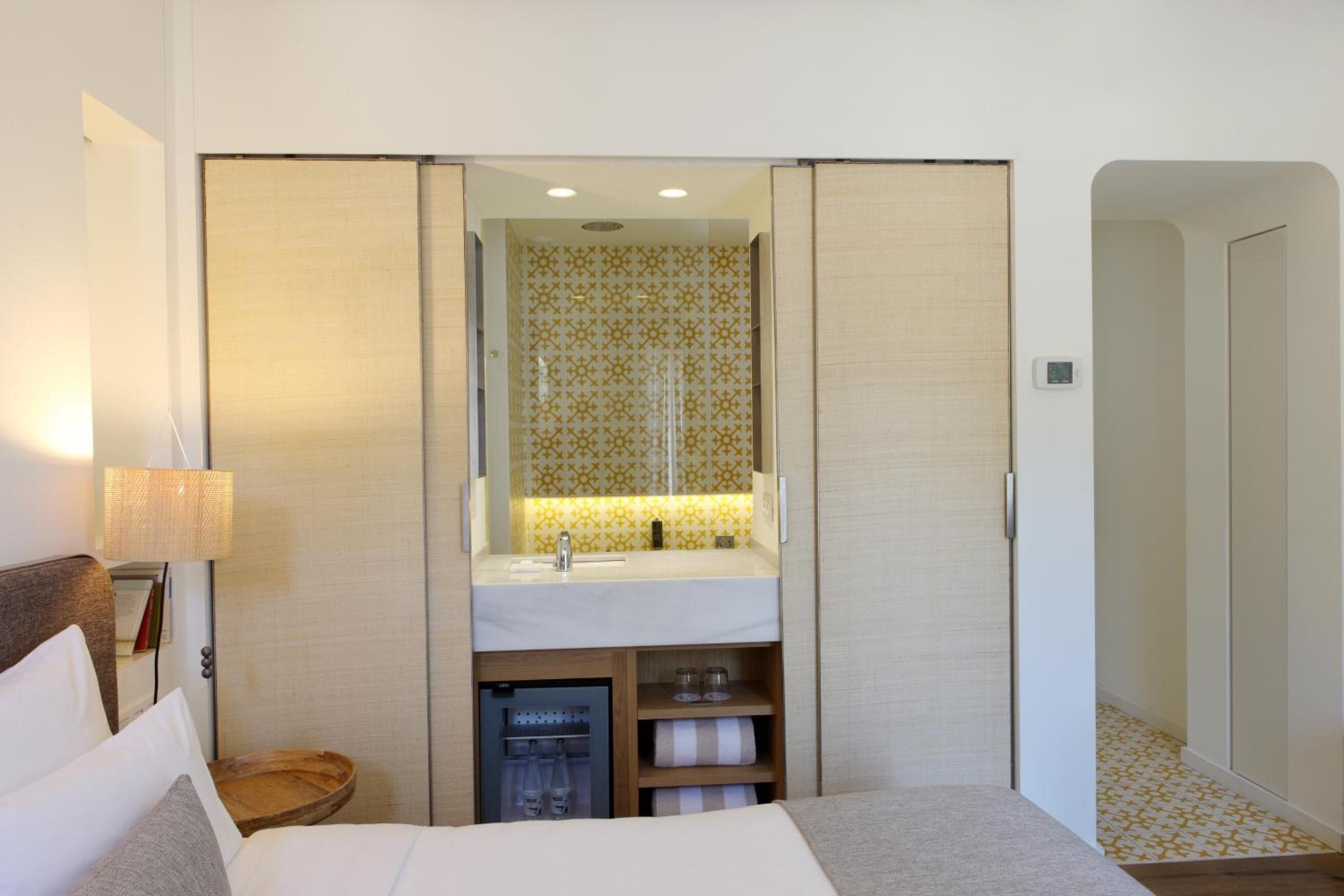 Habitación de hotel con cama, mesita de noche, lámpara, armario y baño con espejo, lavabo y toallero.