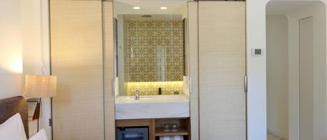 Habitación de hotel con cama, mesita de noche, lámpara, armario y baño con espejo, lavabo y toallero.