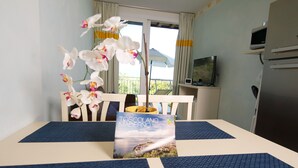 Habitación de hotel con una mesa de comedor cubierta con individuales azules, un libro titulado 'TOSCANO MADERNO' sobre la mesa y orquídeas blancas en un jarrón.