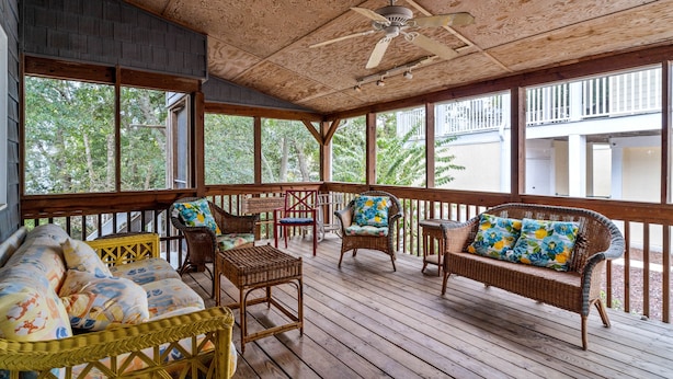 A covered porch with wicker furniture, a ceiling fan, and large windows overlooking a wooded area.