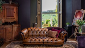 A room with a leather sofa, a cabinet, a painting, and a view of a green landscape. - Fernhill Castle (Keighley)