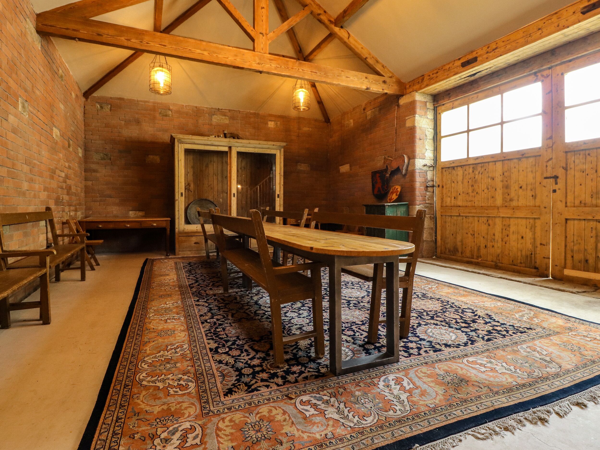 A rustic dining area with a wooden table and benches, a patterned rug, and a brick wall.