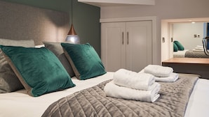 A modern bedroom with a large bed, green and grey pillows, a quilted grey duvet, and a bedside table with folded towels. - Lower Sheriff's Place (Windermere)