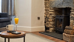 A cozy living room with a stone fireplace, a sofa, a coffee table with a glass of orange juice, and a fireplace log storage. - Lower Sheriff's Place (Windermere)