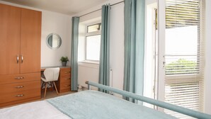 A bedroom with a bed, a wooden wardrobe, a desk with a chair, a window with curtains, and a view of greenery outside. - Delfryd (Y Felinheli)