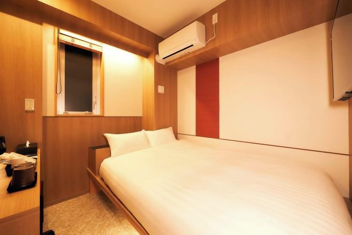 A hotel room with a bed, a bedside table with a lamp, an air conditioning unit, and a window.