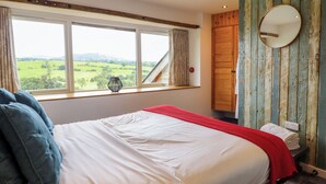 A bedroom with a large bed, a window with a view of the countryside, a wooden wardrobe, and a mirror on the wall.