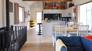 A spacious living area with a high ceiling, wooden beams, a kitchen with modern appliances, and a dining area with white furniture.