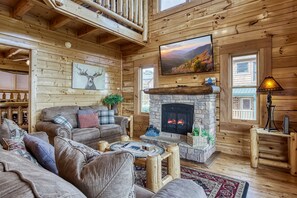 Ein gemütliches Blockhaus-Interieur mit Kamin aus Stein, einem großen an der Wand montierten Fernseher und einer komfortablen Sitzecke mit Sofa und Couchtisch.