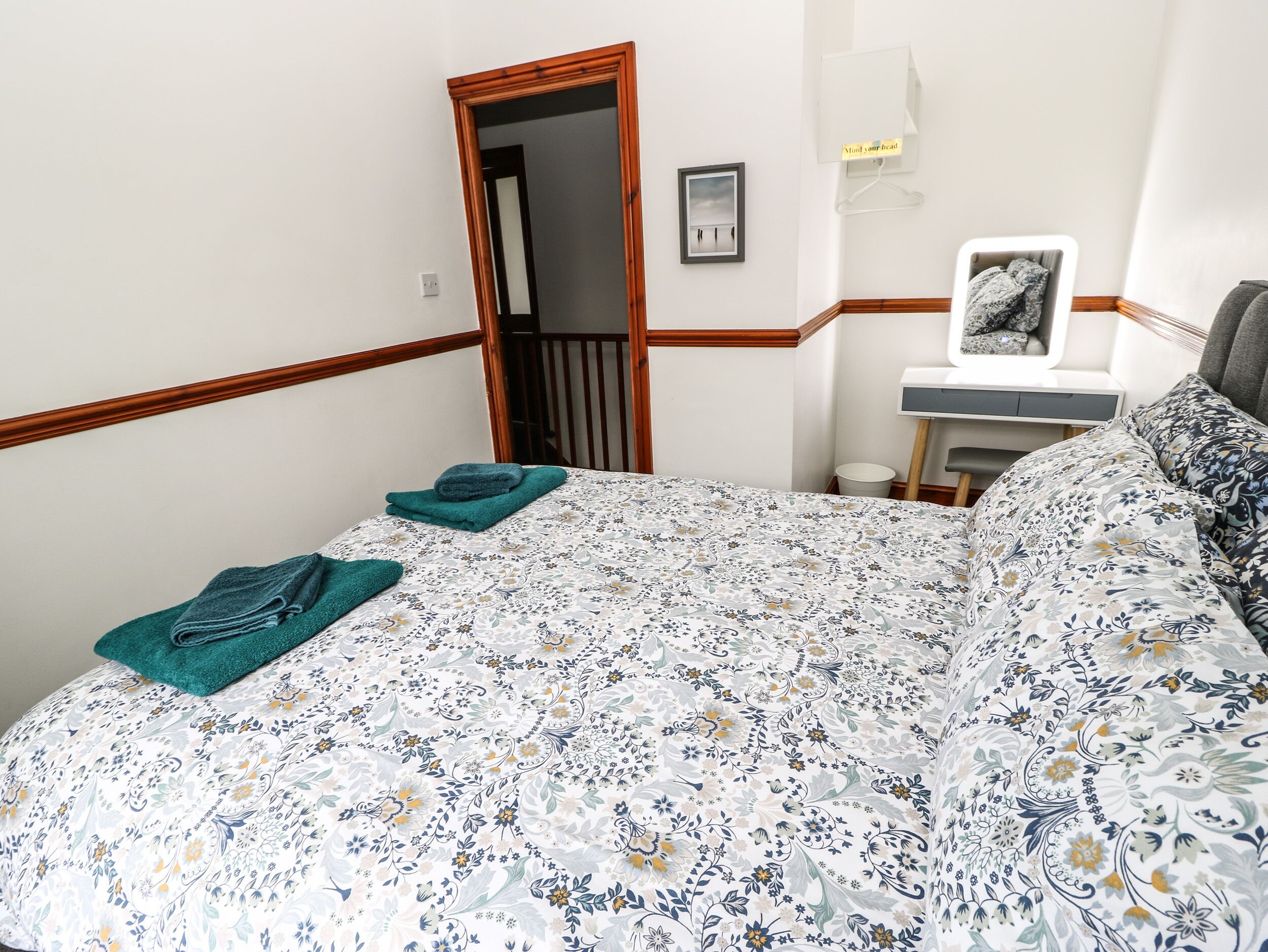 Une chambre avec une couvre-lit à motifs floraux, une tête de lit en bois, une table de chevet avec une lampe et un tableau encadré accroché au mur.