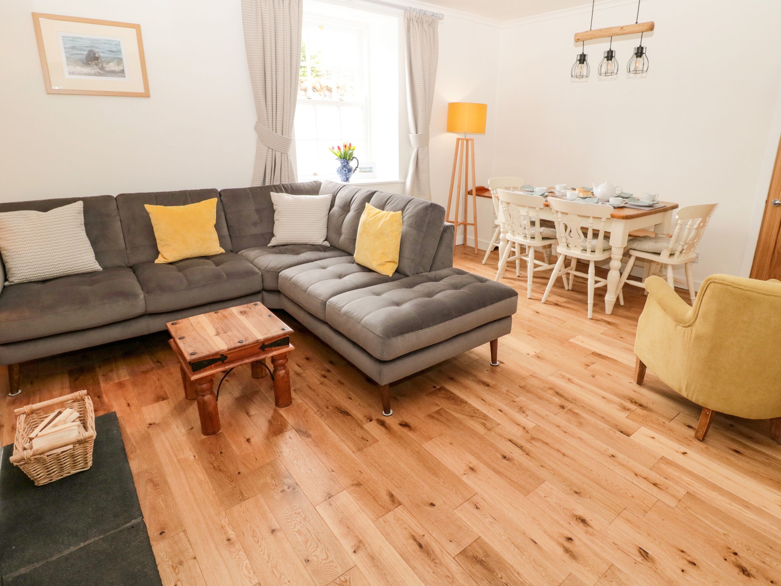 A living room with a grey sectional sofa, a wooden coffee table, and a yellow armchair. There is a dining area with a white table and chairs.