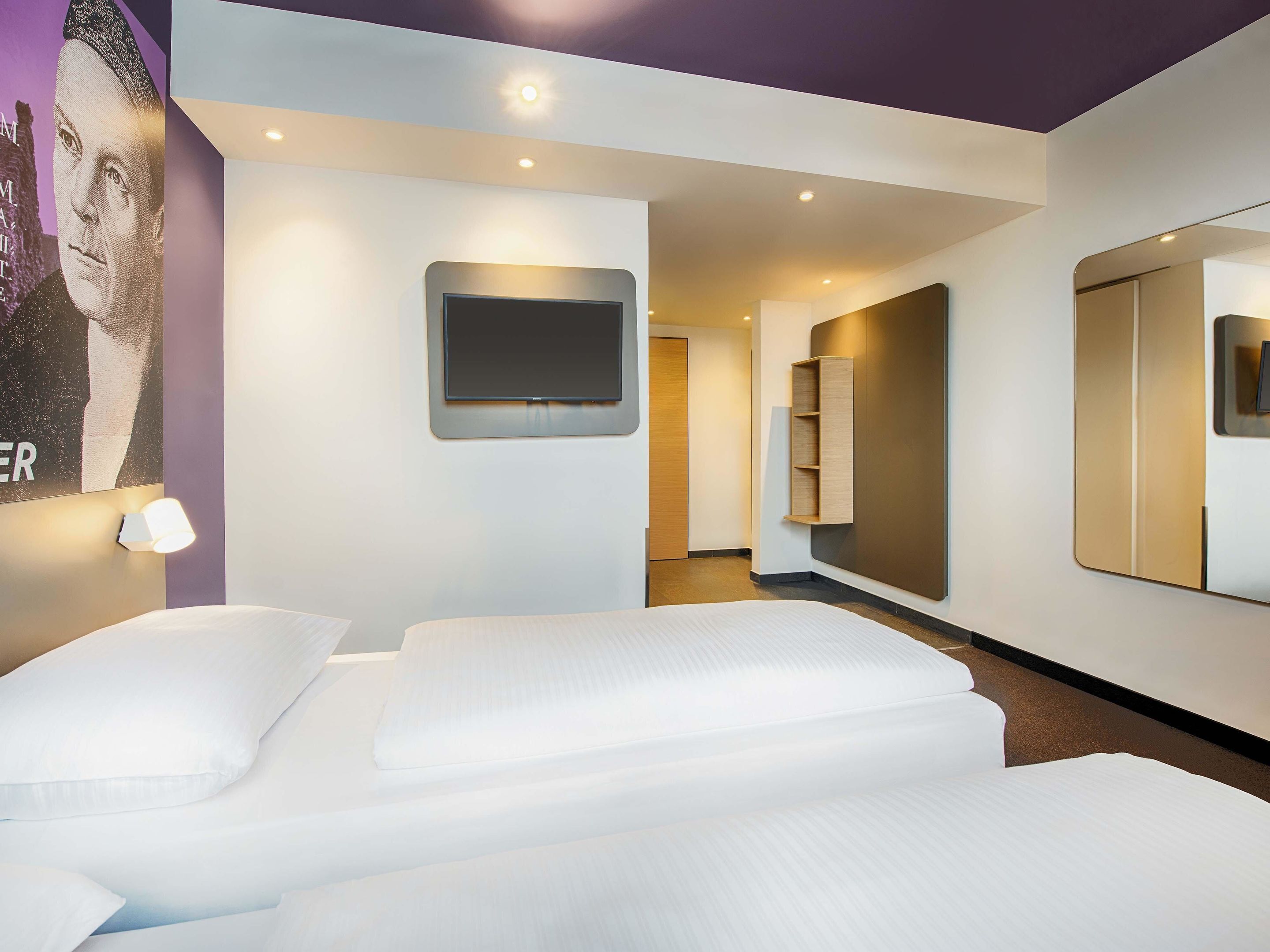 Quarto de hotel moderno com uma cama grande, uma TV de tela plana na parede e um espelho refletindo o ambiente.