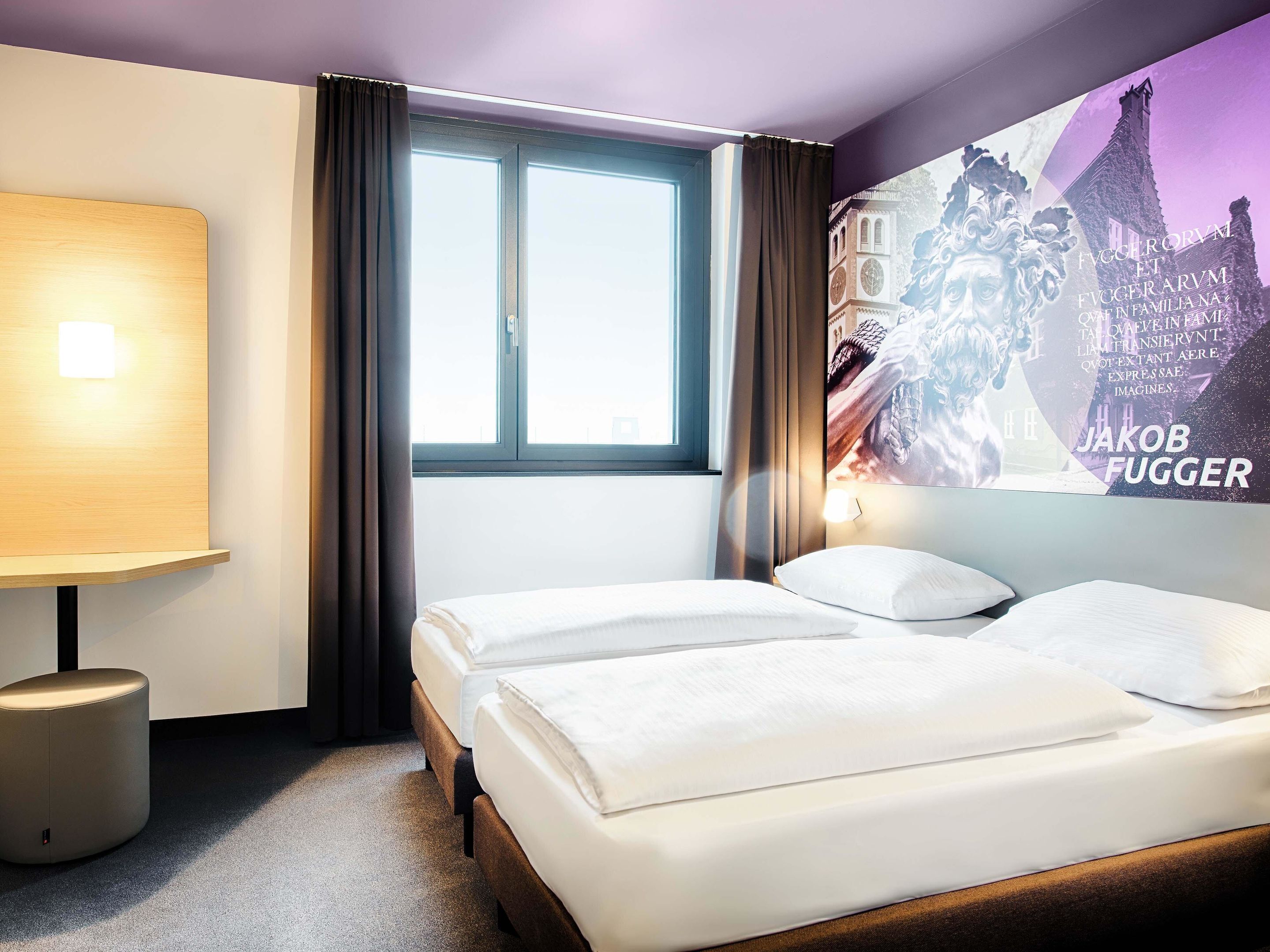Une chambre d’hôtel avec deux lits, une fenêtre avec des rideaux, une lampe fixée au mur et une affiche représentant un bâtiment.