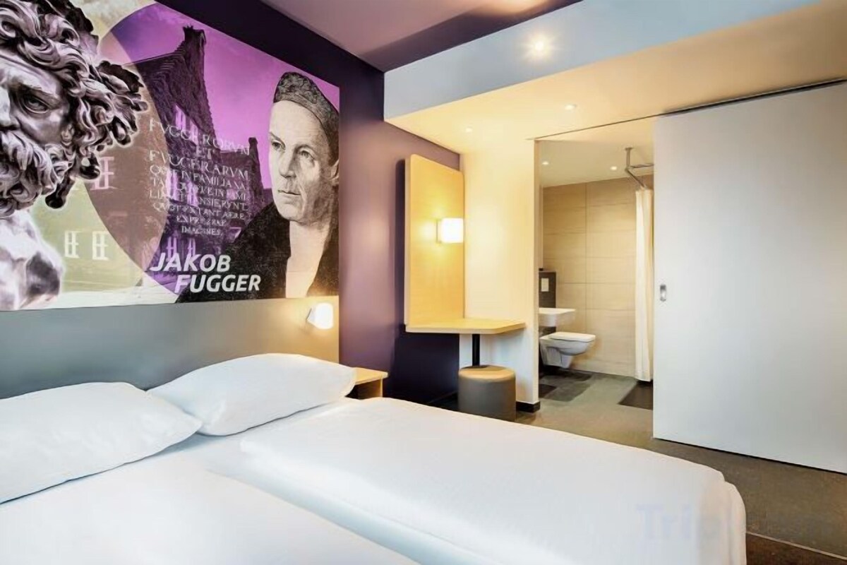 A hotel room with a bed, a bathroom with a toilet and sink, and a wall with a poster.