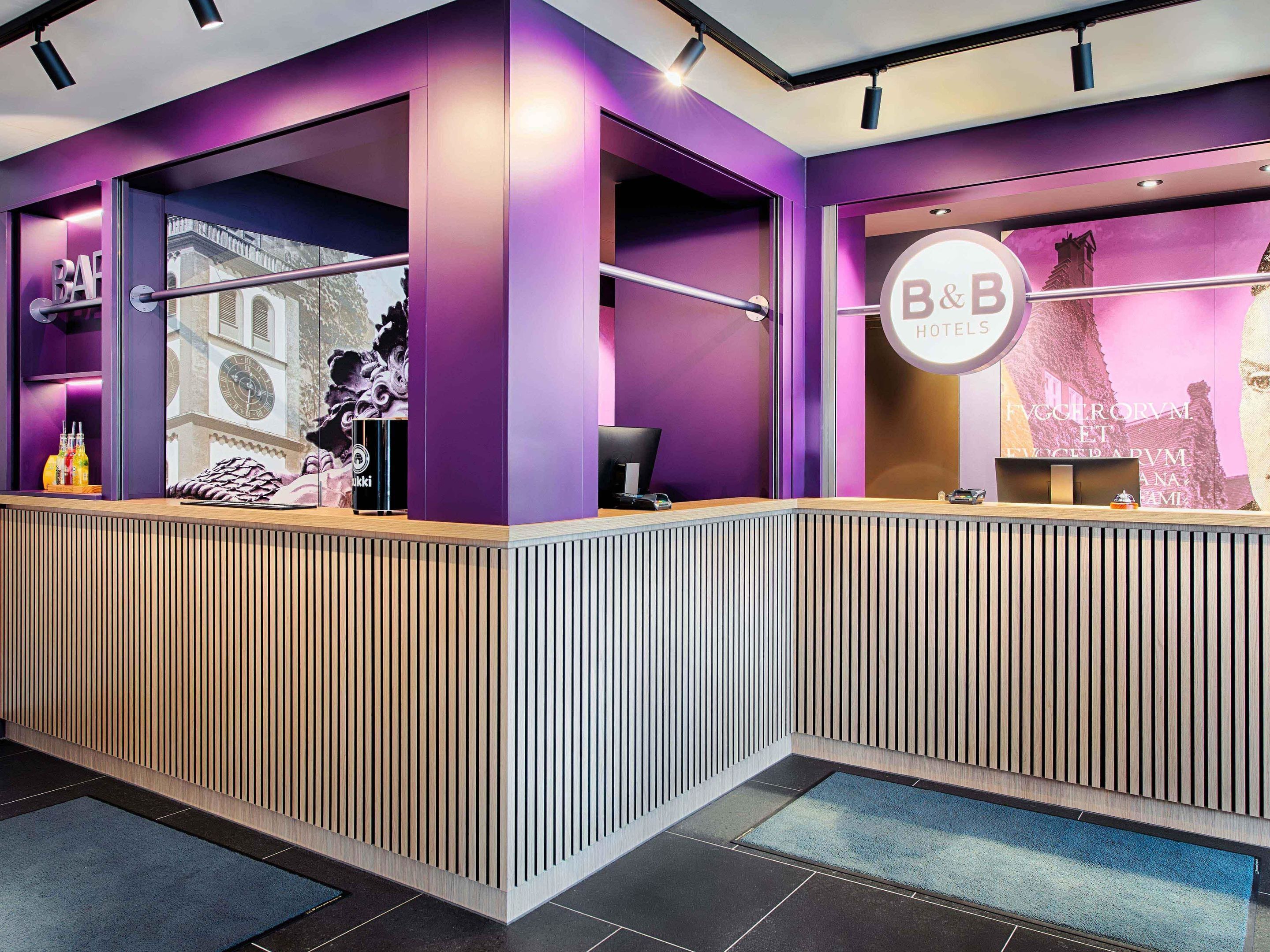 A reception desk with a purple and white color scheme, a large window with a view of a building, and a sign that reads "B&B HOTELS".