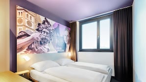 A hotel room with a bed, a desk, a chair, a window with curtains, and a wall with a poster.