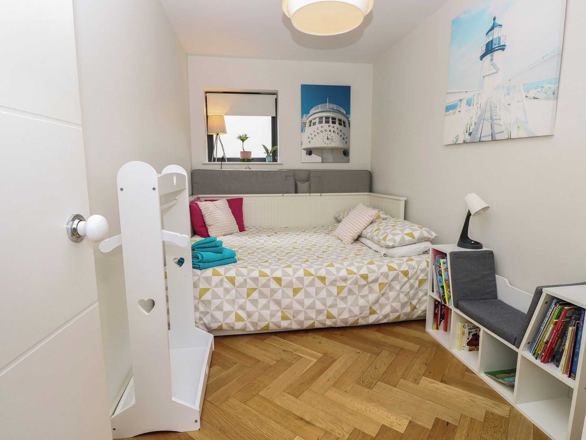 A bedroom with a bunk bed, a bedside table with a lamp, a bookshelf, a window with a view of a lighthouse, and a wall-mounted mirror.