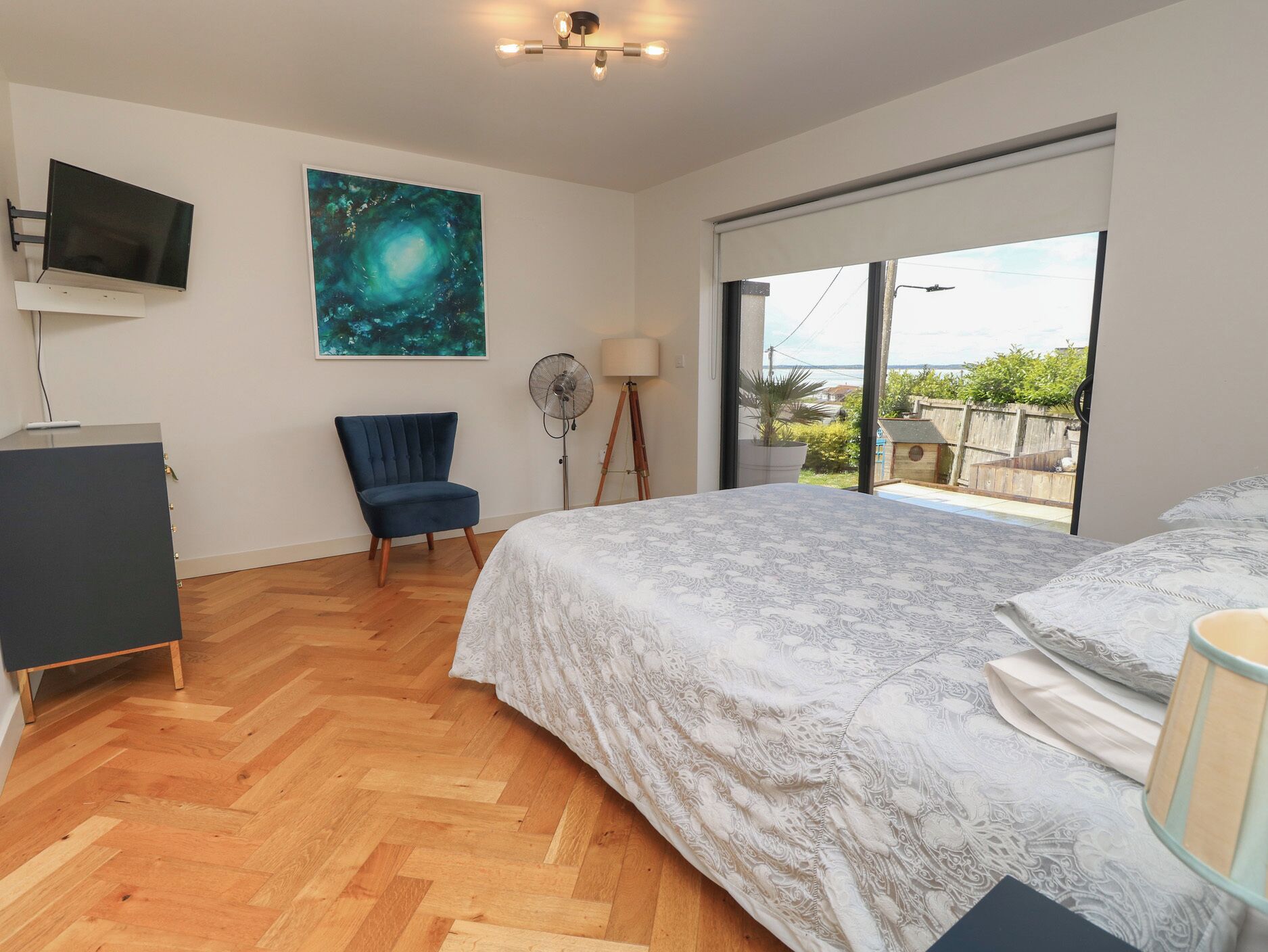 A bedroom with a bed, a chair, a desk, a TV, a fan, and a large window with a view.