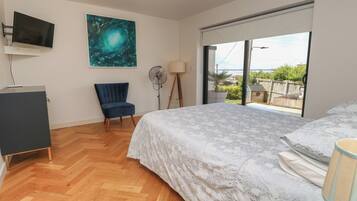 A bedroom with a bed, a chair, a desk, a TV, a fan, and a large window with a view.