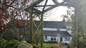 A white house with a tiled roof, surrounded by a garden with a wooden gate and a stone pathway leading to the entrance. - 2 Mellfell View (Penrith)