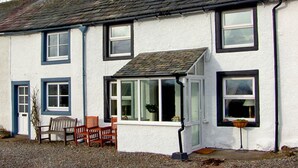 A two-story house with a white exterior and dark trim. There are two visible windows, a door, and a small porch area with a bench and a potted plant. - 2 Mellfell View (Penrith)