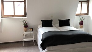 Een slaapkamer met een bed, twee ramen, een kleine tafel met een plant en een witte nachtkastje.