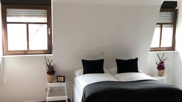Ein Schlafzimmer mit einem Bett, zwei Fenstern, einem kleinen Tisch mit Pflanze und einem weißen Nachttisch.