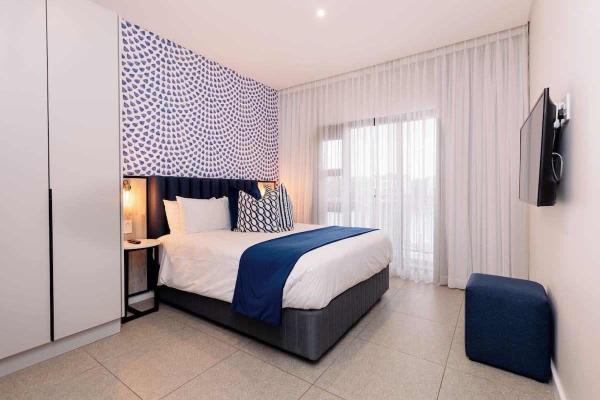 A modern hotel room with a large bed, a bedside table, a TV mounted on the wall, and a window with sheer curtains.