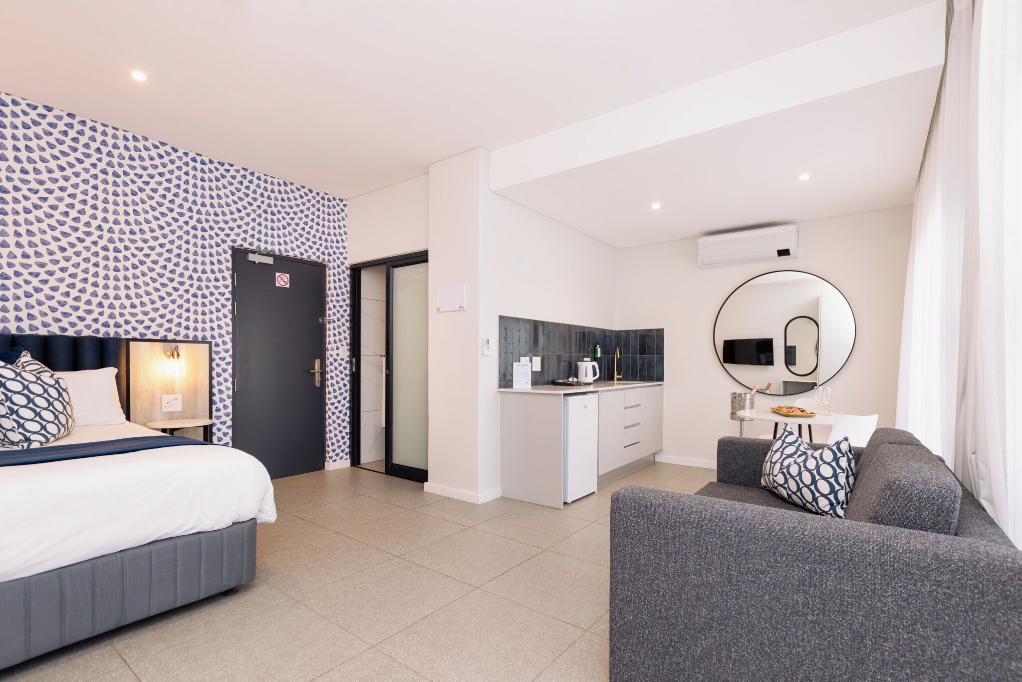 A modern hotel room with a bed, a sofa, a kitchenette, and a bathroom.