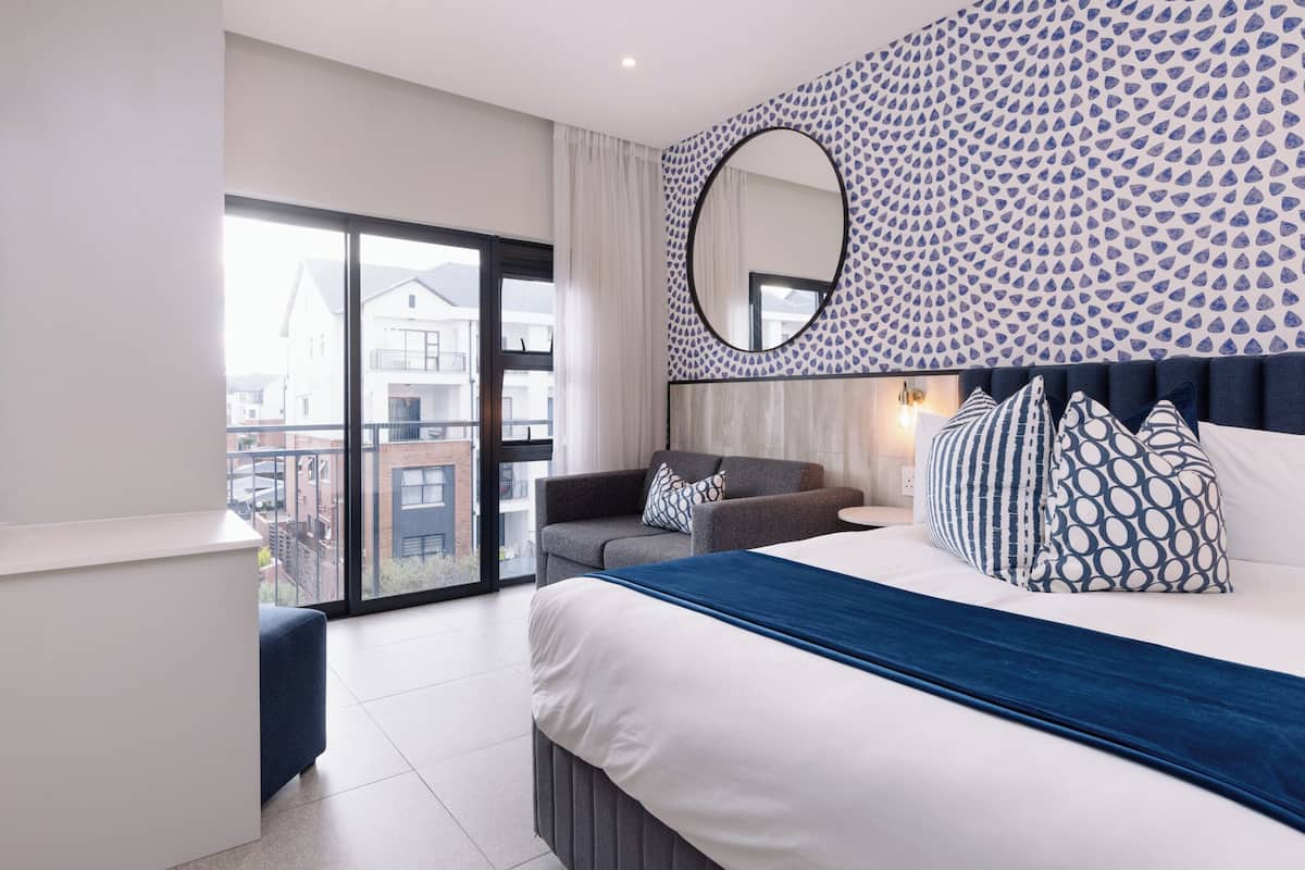 A modern bedroom with a large bed, a sofa, a round mirror, and a patterned wall.