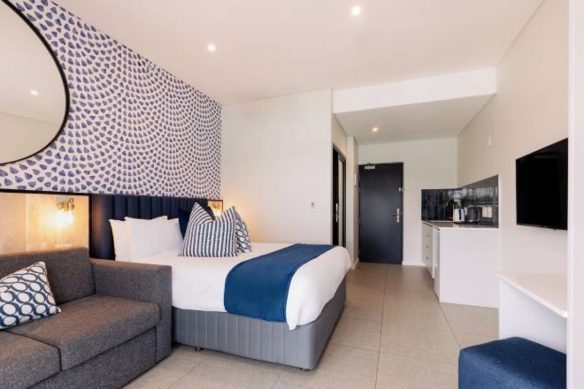A modern hotel room with a bed, a sofa, a dining area, and a kitchen.