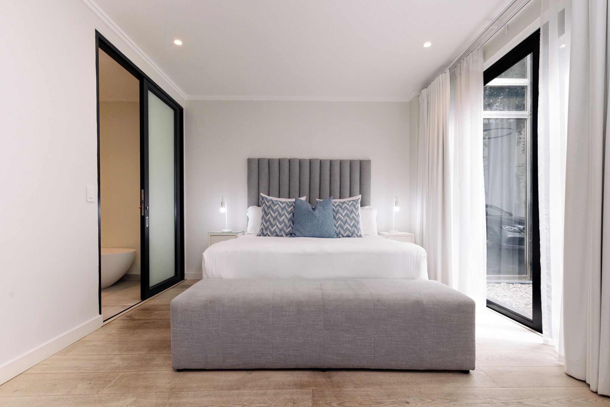 Une chambre moderne avec un grand lit, une tête de lit grise et une porte-fenêtre coulissante donnant sur un balcon.