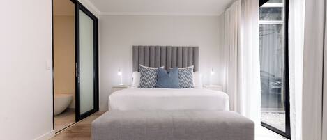 Une chambre moderne avec un grand lit, une tête de lit grise et une porte-fenêtre coulissante donnant sur un balcon.