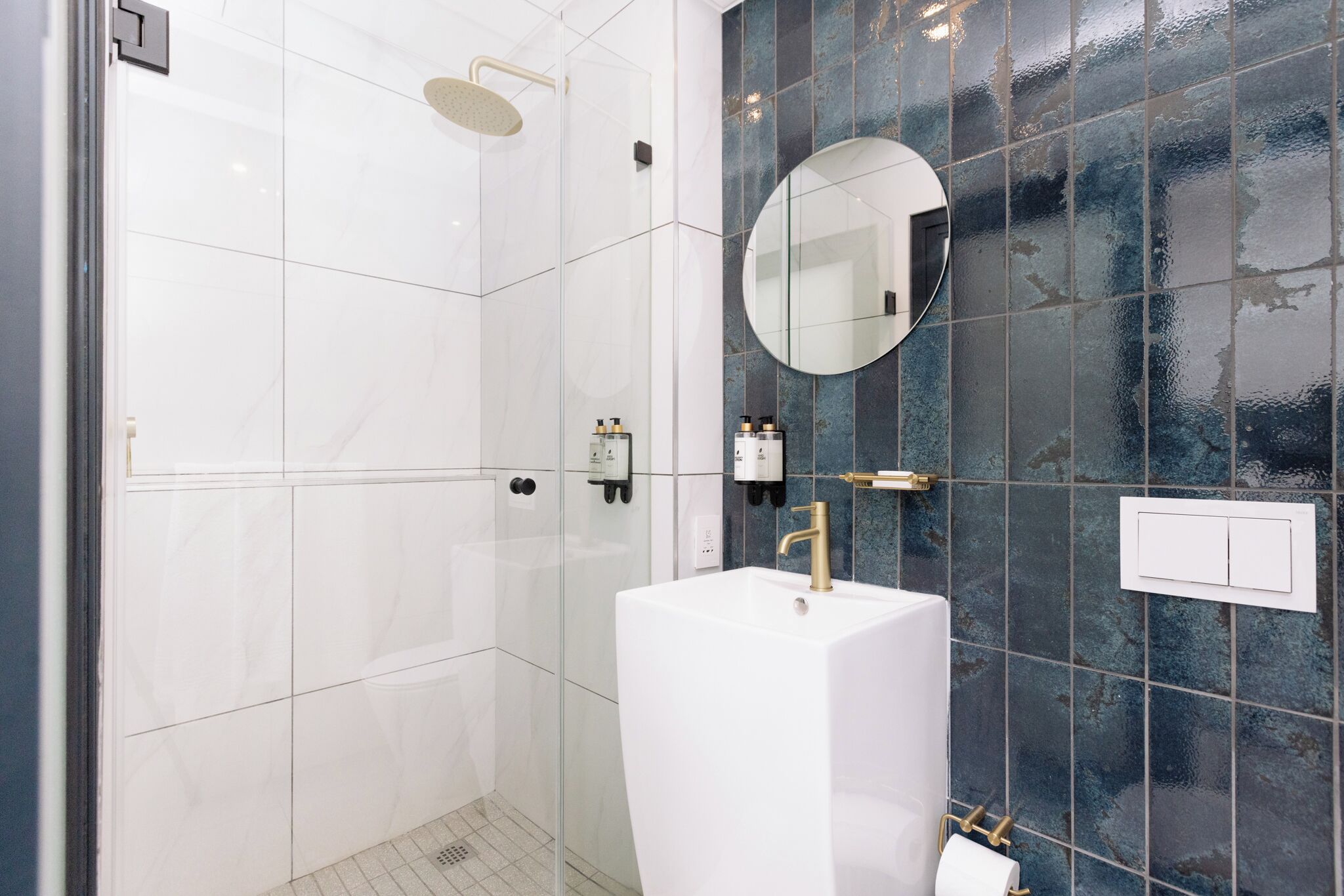 Un baño moderno con cabina de ducha de vidrio, un lavamanos pedestal blanco y un espejo redondo.