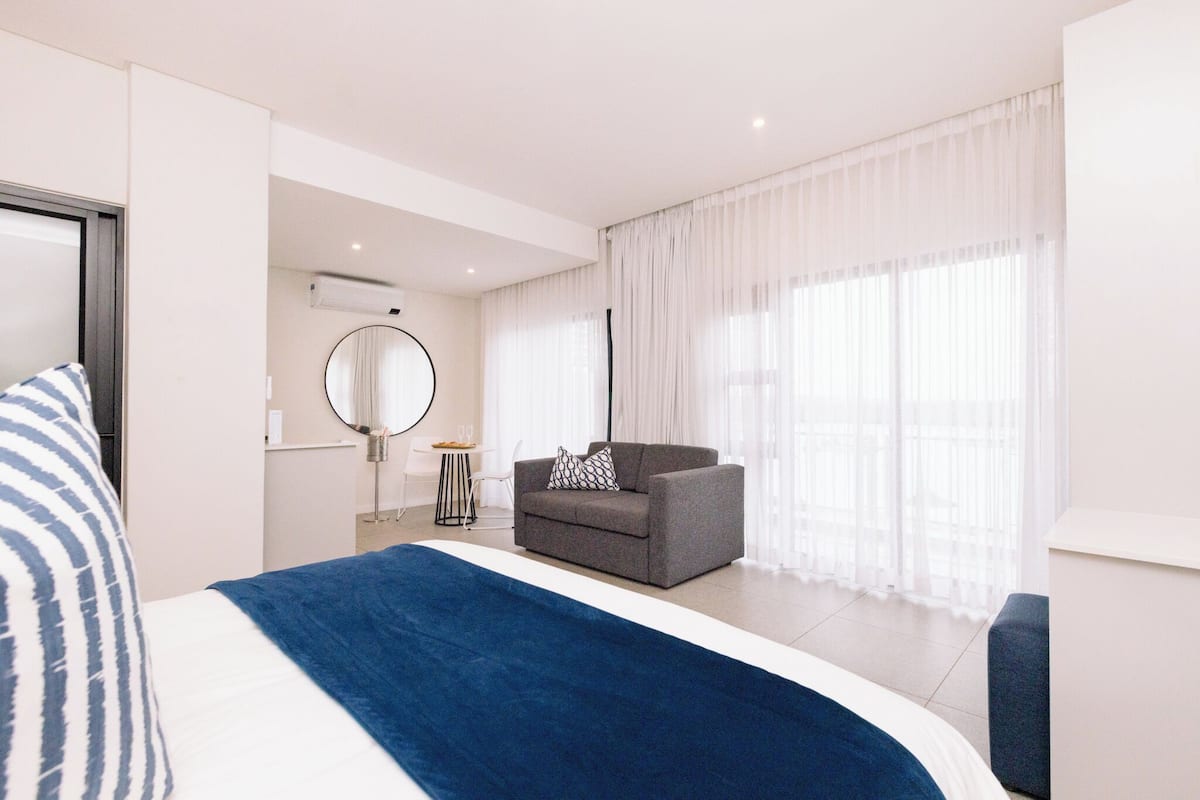 A modern hotel room with a large bed, a grey sofa, a round mirror, and a large window with sheer curtains.