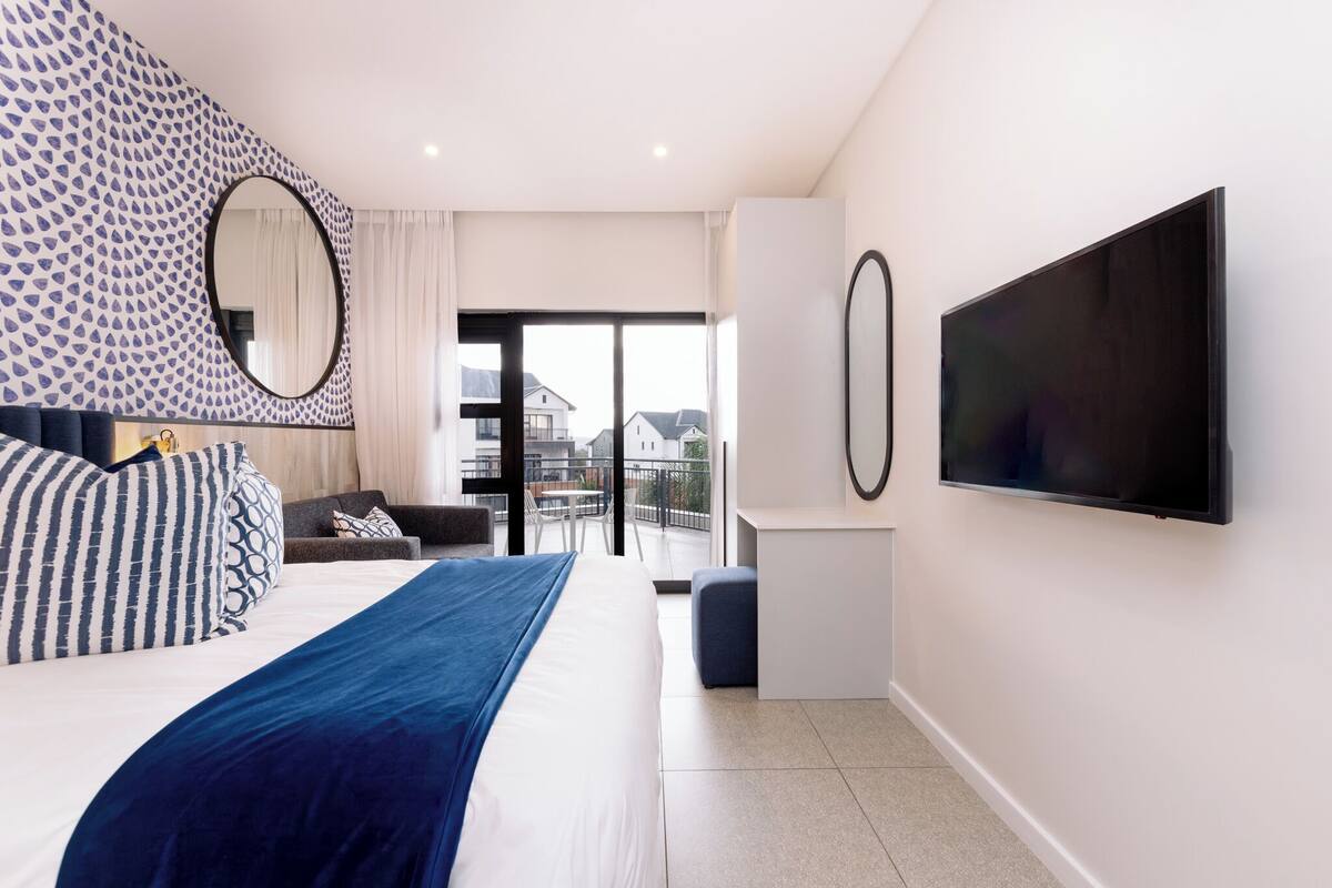 A modern bedroom with a large bed, a TV mounted on the wall, and a balcony with a view of the city.