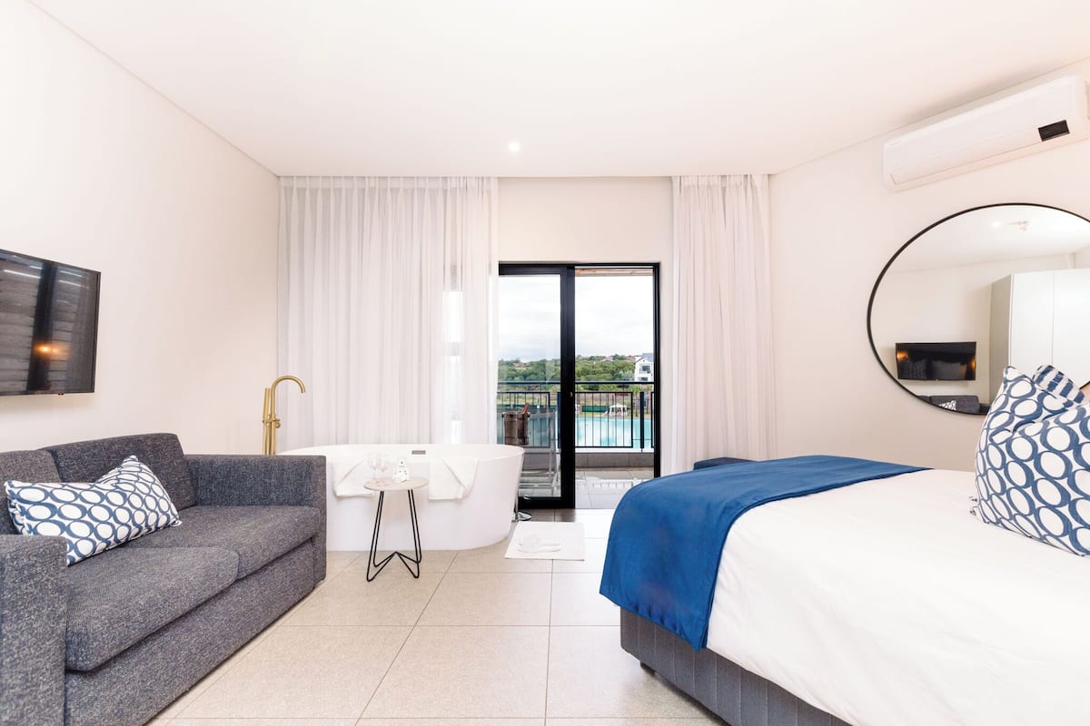 A modern hotel room with a sofa, a bed, a small table, a bathtub, and a balcony with a view.