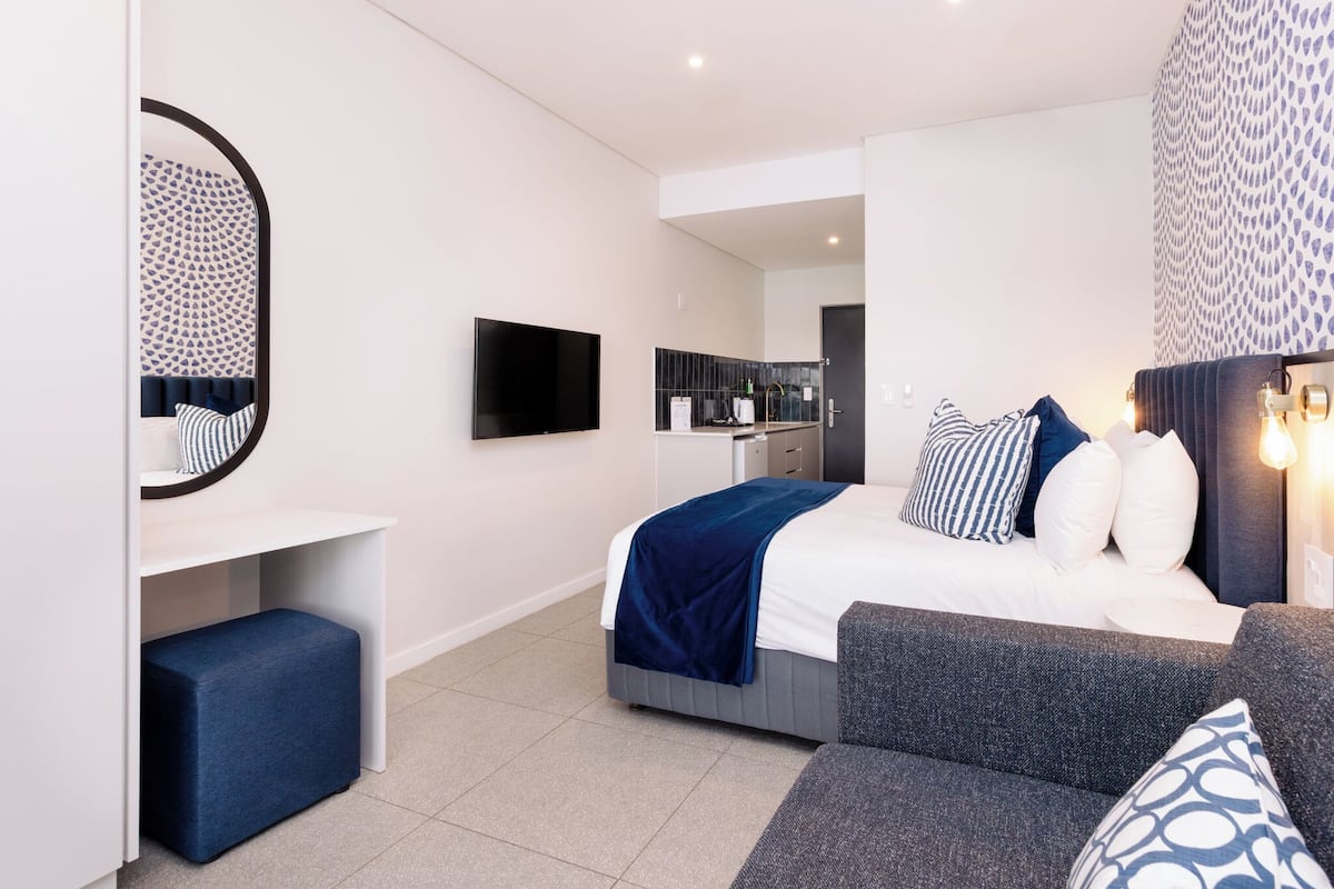 A modern bedroom with a bed, a sofa, a TV, and a small kitchenette.